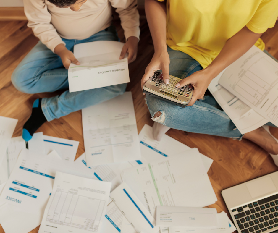 Two people sit on a wooden floor, surrounded by scattered papers and invoices. One holds a calculator, and an open laptop is nearby.