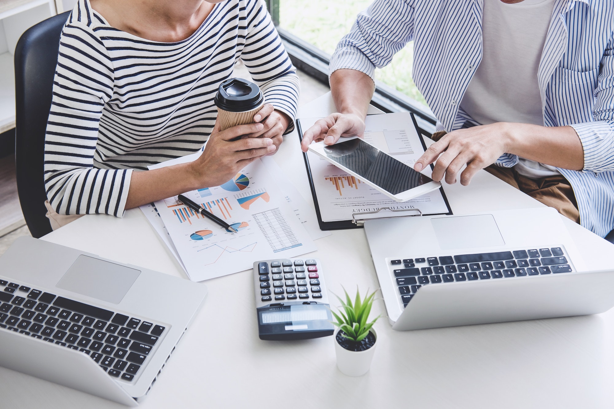 two people sitting Infront of a desk with two laptops on it with one person holding a table and the other holding a cup of coffee