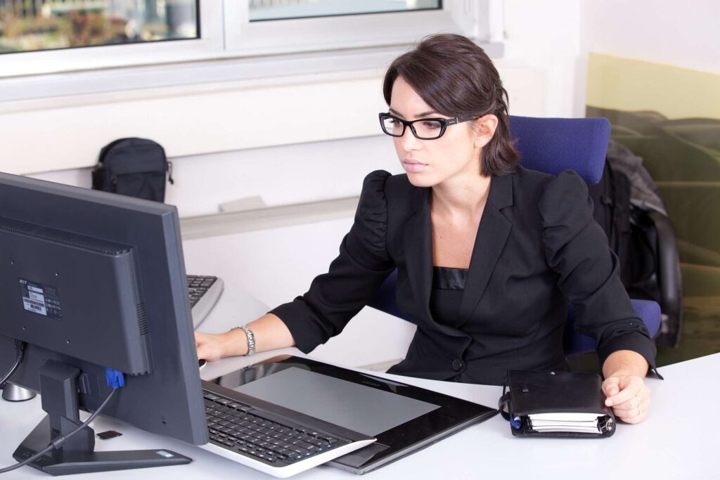 a woman in a black suite wearing glasses sitting Infront a desk working on her computer